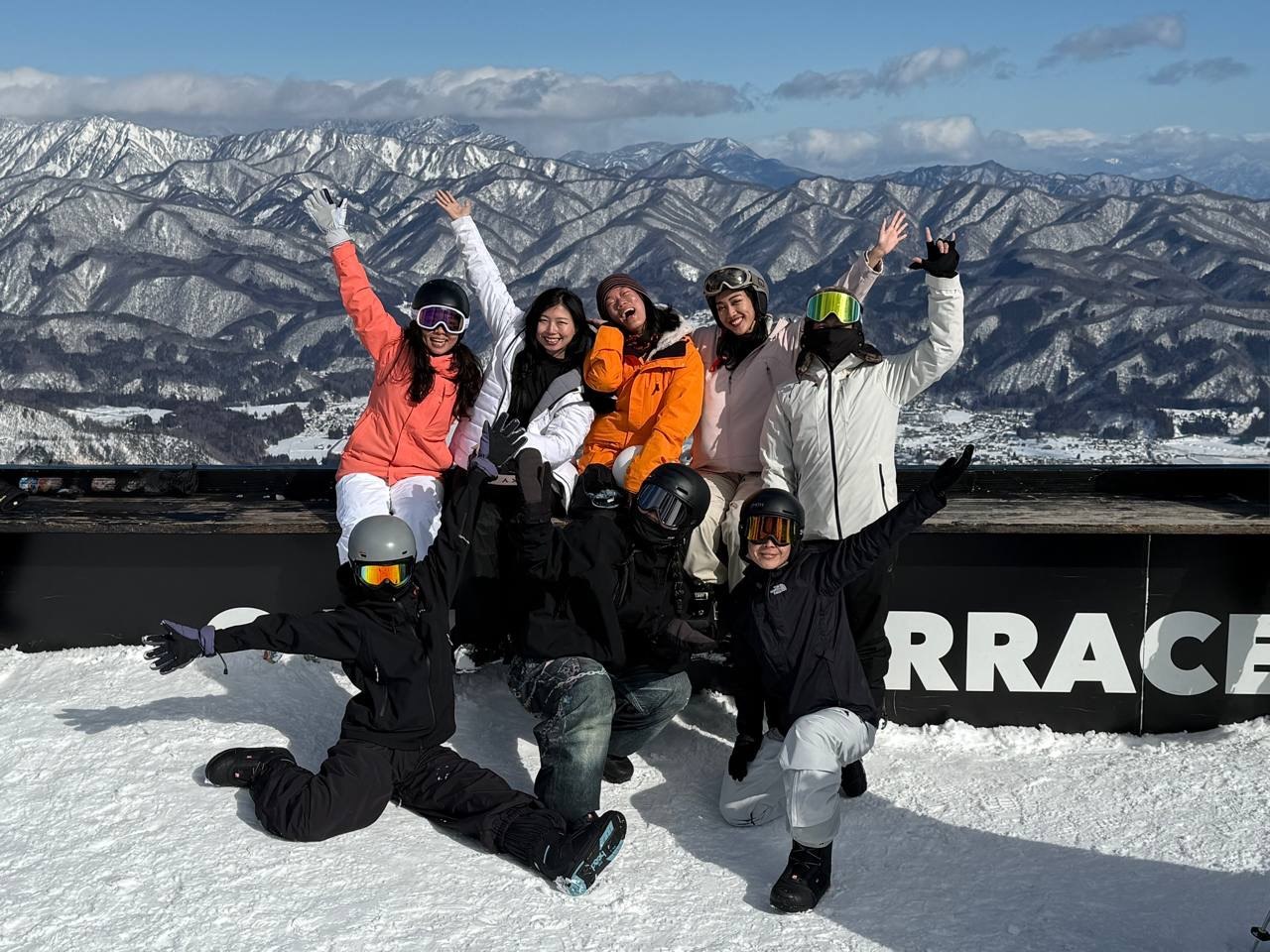 A group of eight travellers in ski gear cheering together on a snow-covered terrace