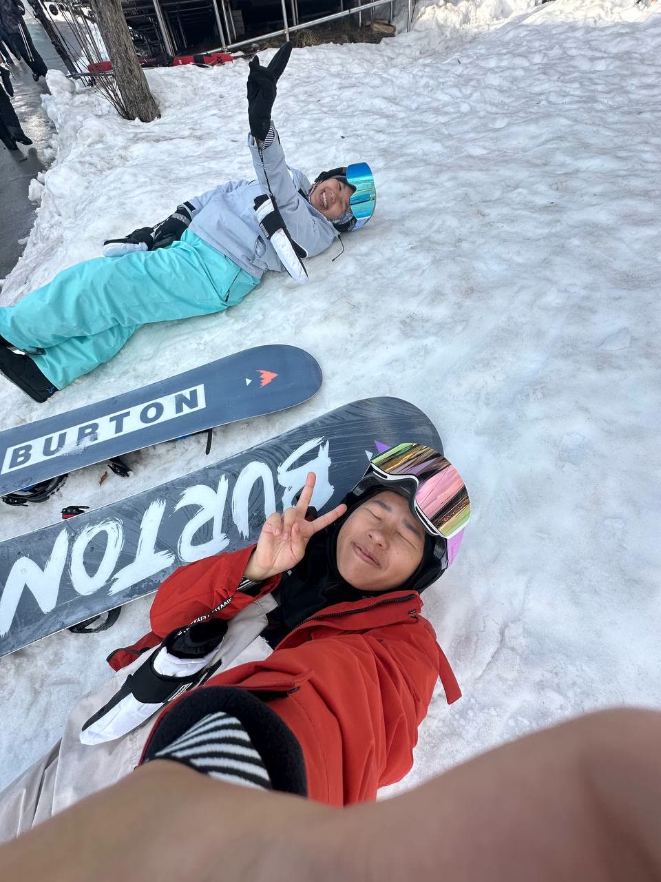 Two women lying in the snow beside their boards, laughing and waving