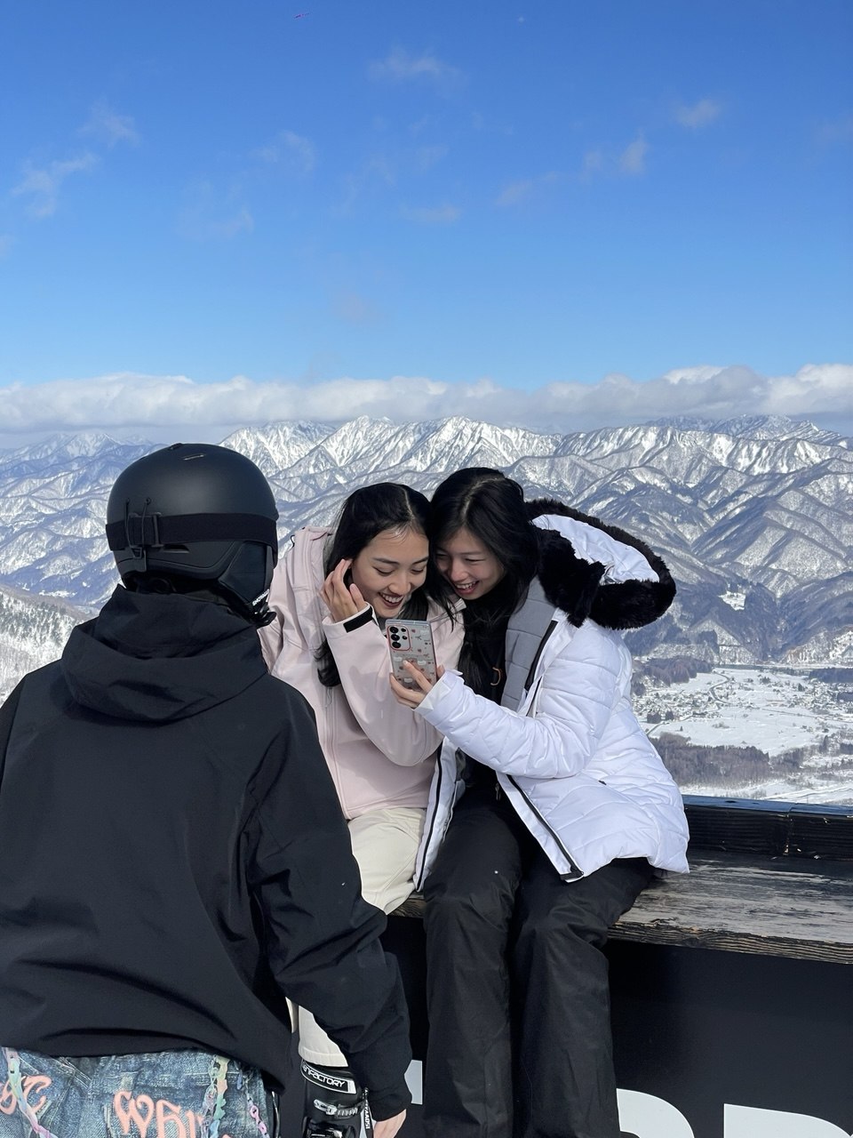 Two travellers laughing over a phone on an alpine deck, snow peaks behind them