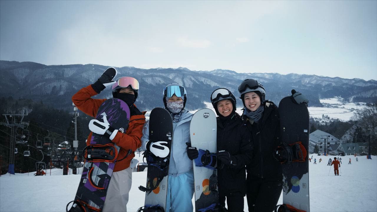 Four women holding their snowboards and smiling on a Hakuba slope, mountains behind them